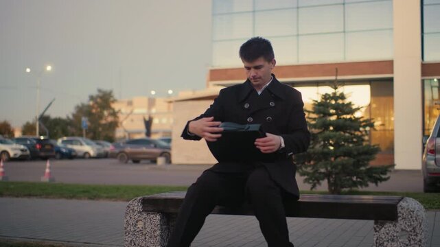 Man in black suit walks up to bench, opens briefcase in outdoor setting with modern building and parked cars, moving car in background at dawn, street light glowing in early morning