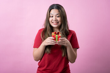 A cheerful young woman with long brown hair and light skin, wearing a red T-shirt, holds a small red gift box with a green ribbon and a decorative bow, smiling brightly against a pink background.