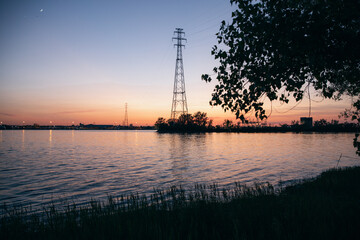 view of a tall electrical tower as seen from the other side of a river on a sunset with a tree in the foreground