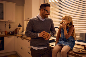 Happy little girl eating sandwich while talking to her father in kitchen.