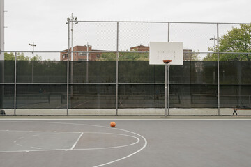 outdoor concrete basketball court in summer with a tall metal fence on a cloudy day with a ball on the ground