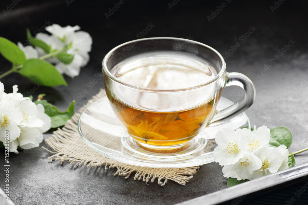 Wall mural Jasmine herbal tea in a glass cup with flowers on black background, closeup, spring floral drink, natural medicine and naturopathy concept