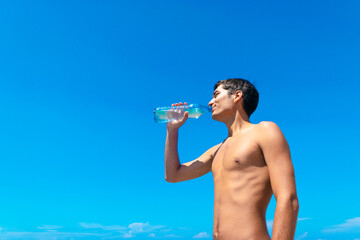 Young tanned smiling guy drinks water from a bottle against a blue cloudless sky.