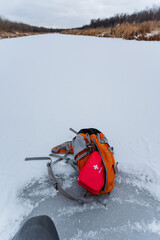 A backpack is casually sprawled on the smooth icy surface, right next to a gently flowing river, creating an intriguing scene amidst the beautiful snowy winter landscape surrounding it