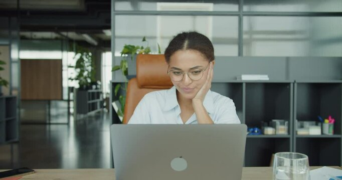 A focused young woman in glasses sitting at a modern office desk, looking at her laptop screen with a thoughtful expression in a professional and organized workspace