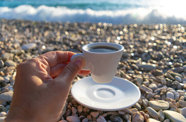 Woman's hand holds a white cup filled with coffee. Pebble beach with sea foam, illuminated by the...