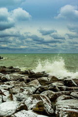 Waves crash onto ice covered rocks with textured blue clouds in the background room for text vertical image shot on Kew beach in Toronto suitable as backdrop or banner