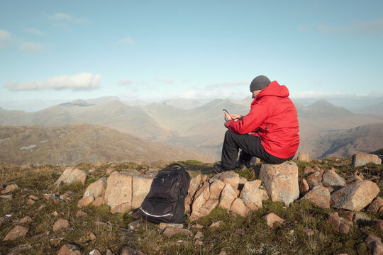 Hiker man sitting on the big stone on the top of the mountain and using his phone at the background of morning landscape with mountains. View from The Devils Staircase to Binnein Mor. Scotland