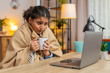 Sick ill Indian young woman freelancer working on laptop wrapped in plaid sit shivering from cold...