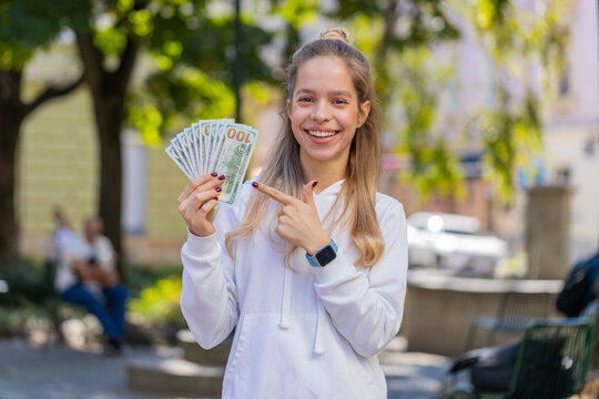 Portrait of successful rich Caucasian girl gambler gambling addiction business woman winner wins days holding fan of dollars, shows finances with thumbs up gesture receives loan salary on city street.