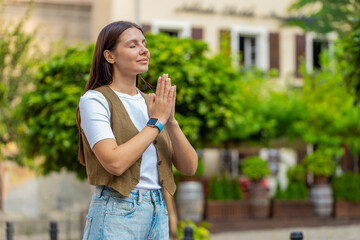 Fototapeta premium Young European woman praying with closed eyes to god asking for blessing, help, forgiveness outdoors. Pretty girl clasping hands wishing luck in sunny urban city street. Town religion lifestyles.