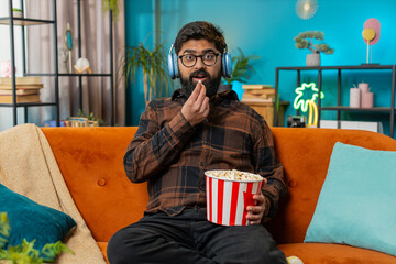 Relaxed Indian young man eating popcorn and watching movie wow expression sitting on sofa in living...