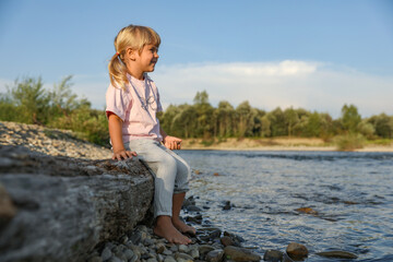 Cute little girl sitting on tree trunk near river. Child enjoying beautiful nature