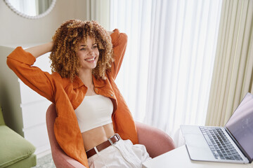 Young woman with curly hair wearing casual outfit, smiling while working on laptop at home Cozy...