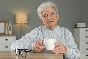 Sad senior woman feeling lonely at wooden table with cup and glasses indoors