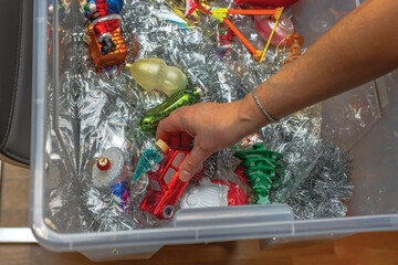 Hand reaching into plastic storage box filled with colorful vintage Christmas ornaments and shiny silver tinsel decorations.