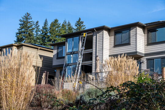 Workman high up on an aluminum extension ladder against a large two-story house, window replacement construction project, outside on a sunny winter day
