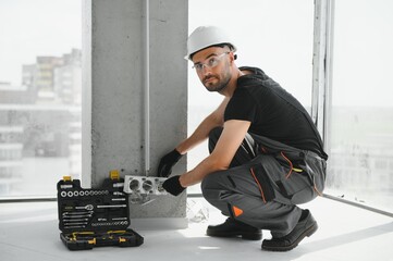 Confident professional electrician in uniform using screwdriver while replacing a socket in...