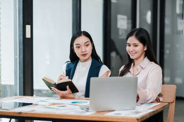 Two businesswomen discussing ideas in a contemporary office, using a laptop and taking notes.