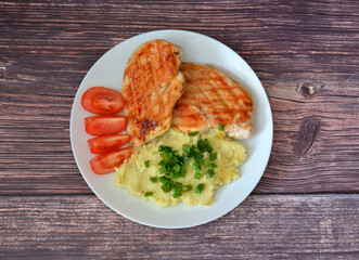 A plate with two pieces of grilled chicken fillet with tomatoes and mashed potatoes with green onions on a wooden table.