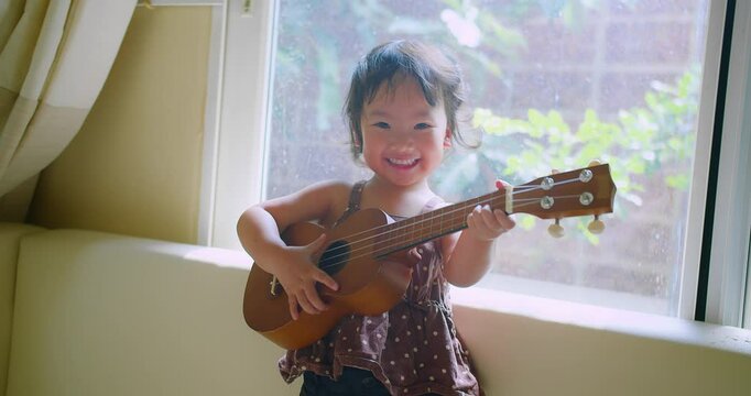 Happy young Adorable Asian Child joyfully Playing Ukulele by a Sunlit Window with Greenery in the Background, Capturing a Joyful and Musical Moment Indoors