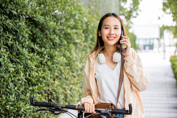 Fototapeta premium Asian young woman walking in countryside outdoor with her bicycle she using mobile phone talking with friend, Happy female stand on city street with bike using smartphone for summer sightseeing walk