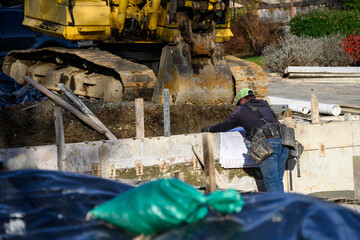 Job foreman with toolbelt looking over house architectural drawings for construction project in foundation concrete pouring stage, home construction on a sunny winter day
