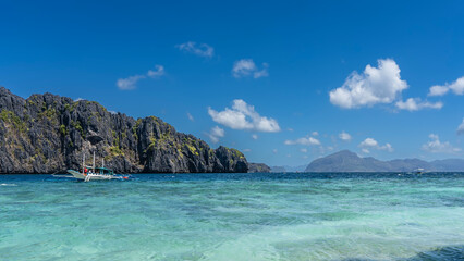 The traditional Filipino bangka boat is anchored in the aquamarine ocean. Sheer karst cliffs against a blue sky and clouds. Green vegetation on steep slopes. Philippines. Palawan. Bacuit bay
