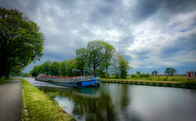 Fototapeta premium A charming canal scene with a slim barge navigating lush greenery and dramatic clouds, creating an aweinspiring vibe