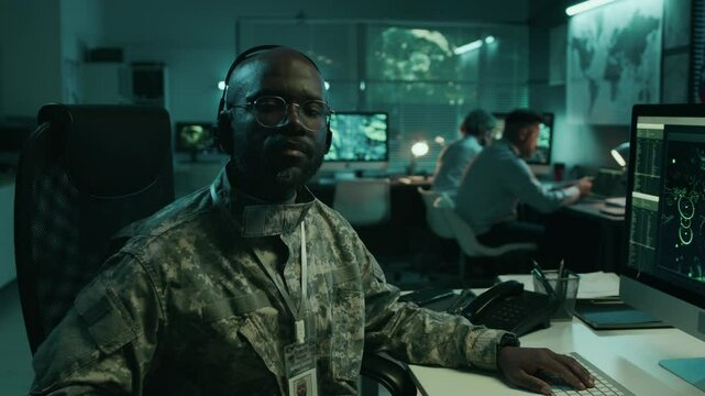 Medium closeup portrait of African American male satellite surveillance operator in camouflage uniform sitting at at computer at military operations center, looking at camera, in front of USA flag
