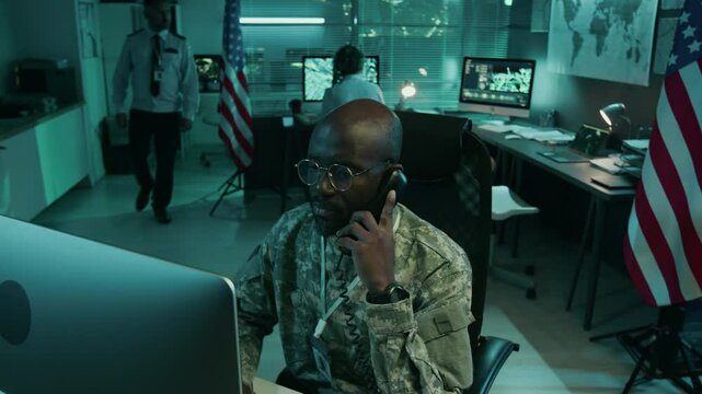 Medium high-angle shot of African American male military officer in camouflage uniform working on computer in command center with USA flag and Caucasian colleagues, talking on landline phone