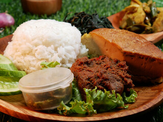 Closeup Padang Rice is a complete traditional food from Padang, Indonesia on a wooden plate, containing white rice, beef rendang, omelette, sliced cucumber, green chili sauce and jackfruit vegetable.