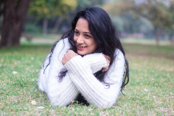 Young happy indian woman wearing warm white sweater lying outdoor at park in winter season.