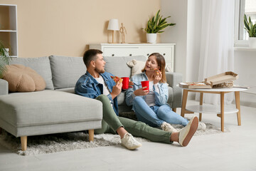 Obraz premium Beautiful young happy couple with tasty pizza pieces and cups of soda sitting on floor at home