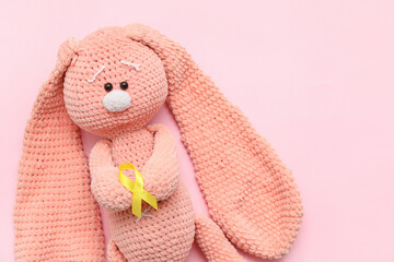 Knitted toy bunny and golden awareness ribbon on pink background, closeup. International Childhood Cancer Day