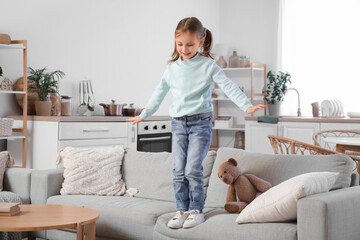 Cute little girl standing on edge of sofa at home. Child in danger