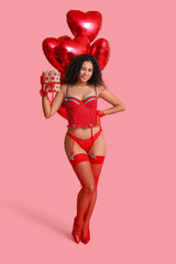 Beautiful young African-American woman in stylish red lingerie with gift box and air balloons in shape of heart on pink background. Valentine's Day celebration