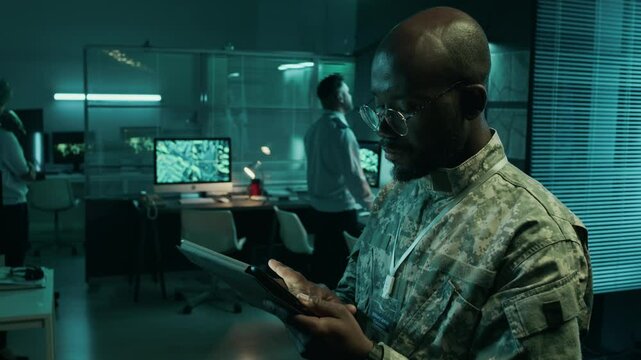 Medium side shot of African American army field commander in camouflage uniform standing in operations center, using tablet computer with digital satellite maps to plan advance or enemy attack