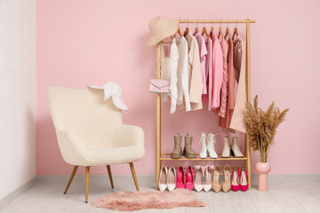 Rack with clothes, shoes and white armchair near pink wall in room