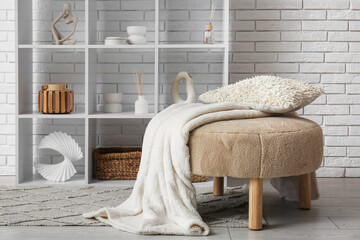 Beige pouf with cushion, plaid and shelving unit near white brick wall in room