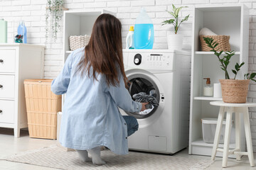 Young woman putting clothes into washing machine in laundry room, back view