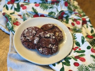 Freshly homemade red velvet crinkle Christmas cookies sitting atop a plain stoneware plate, on top of a wooden table with festive holiday linens.