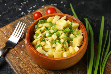 Wooden bowl of tasty potato salad with green onion and tomatoes on black background
