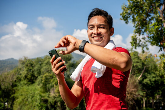 A happy Asian man in sportswear checks his calorie count on his smartwatch while jogging in a park.