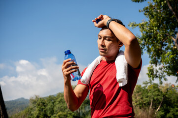 A sweaty, tired Asian man in sportswear drinks water while running in a park on a hot, sunny day.