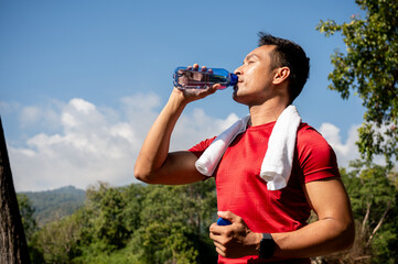 A sporty Asian man drinks water from a bottle, feeling refreshed after his workout in a park.