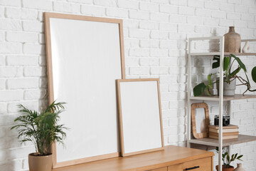 Chest of drawers with houseplant and blank posters near white brick wall in room