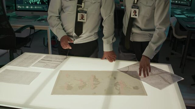 Tilt-down medium closeup of male and female Caucasian military officers in uniform with ID badges and shoulder patches discussing maps and intelligence documents on light table in operations center