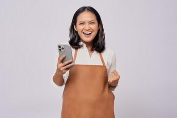 Smiling young Asian woman barista barman employee wearing a brown apron working in coffee shop holding mobile phone and celebrating success isolated on white background. Small business startup concept