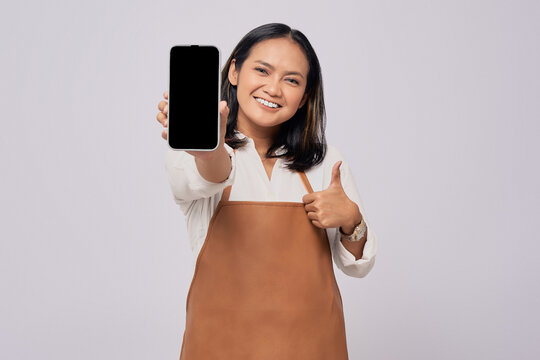 Smiling young Asian woman barista barman employee wearing brown apron working in coffee shop showing mobile phone blank screen mockup and thumb up sign isolated on white background